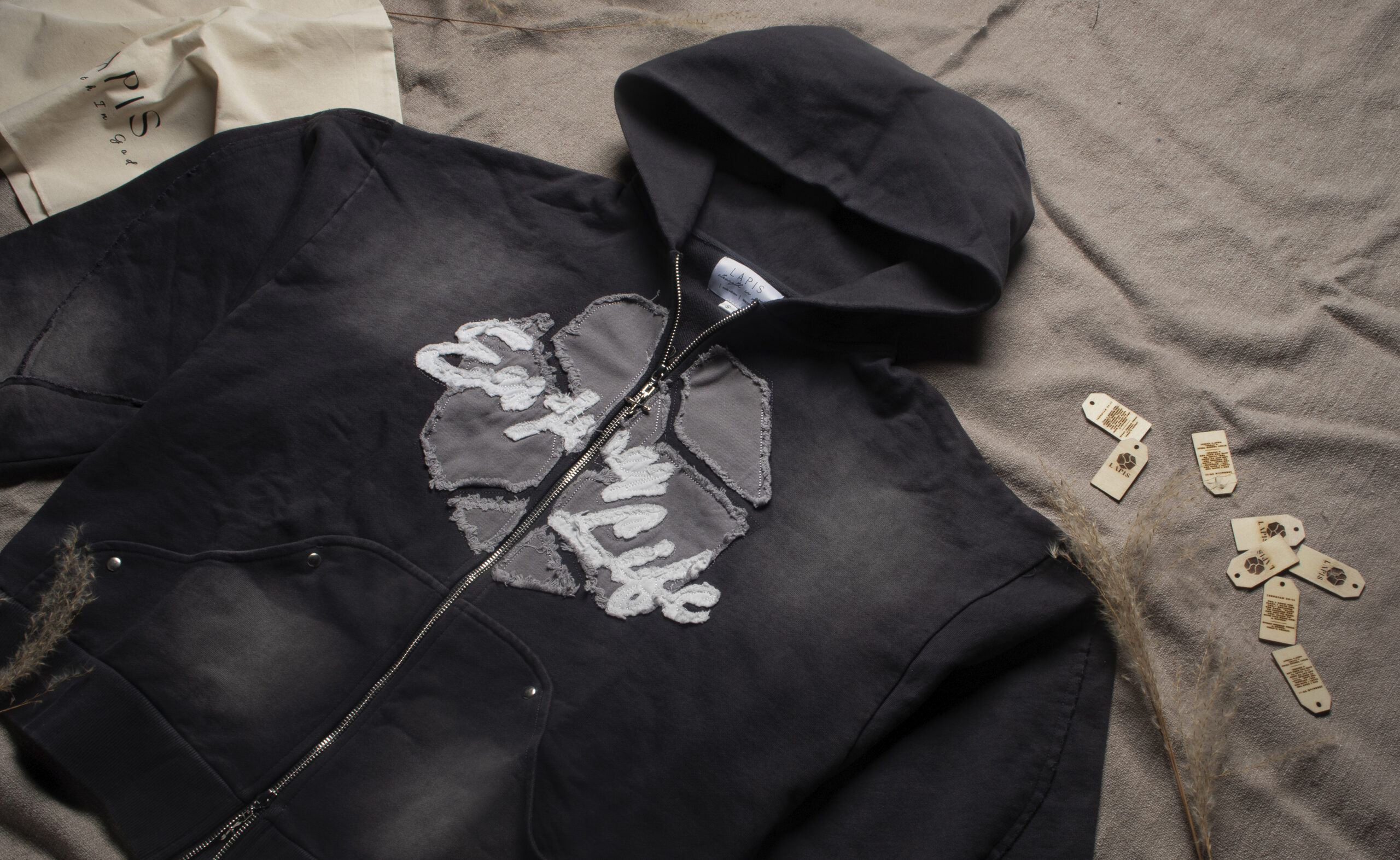 A grey 'Enjoy Life' hoodie laid out on the ground, with wooden tags at the top right and a Lapis bag positioned at the top left.