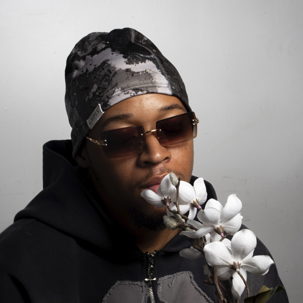 A headshot of a man wearing a grey 'Enjoy Life' hoodie, camo beanie, and sunglasses as he admires a white flower, captured in a calm and reflective moment.