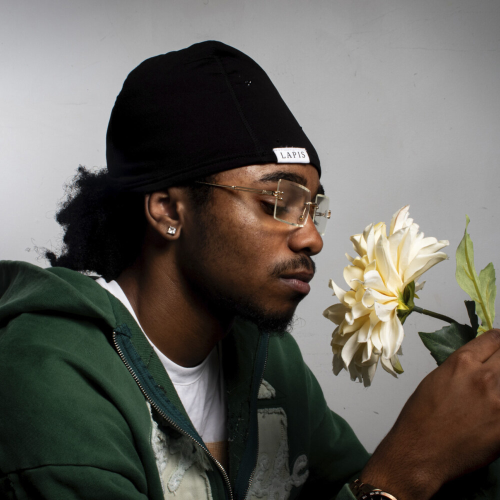 A man wearing a green hoodie and a black hat admires a yellow flower, capturing a quiet and thoughtful moment.