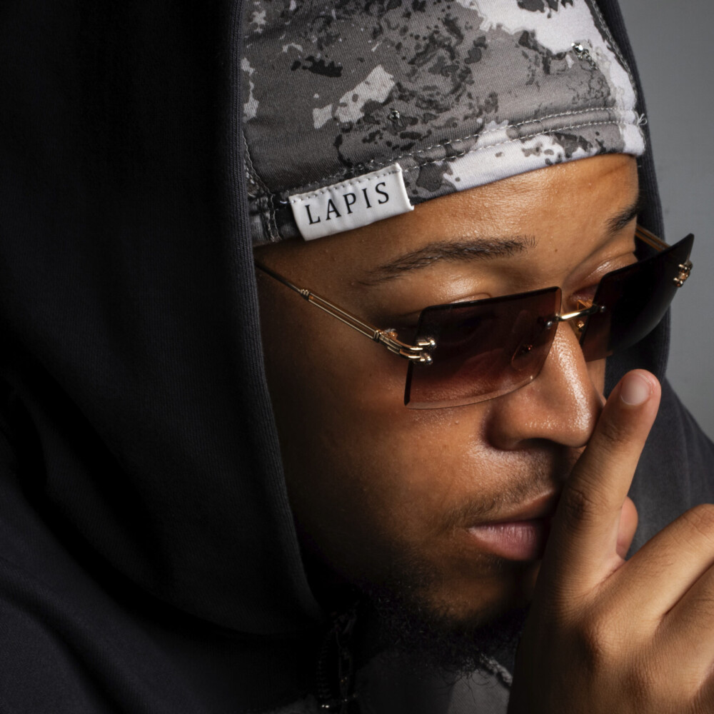Man wearing a hooded sweatshirt, grey camouflage beanie, and dark sunglasses, facing slightly right with a serious expression.